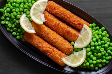Homemade breaded cod fish fingers with garden peas and sliced lemon in a black stoneware dish. On a black wood background.