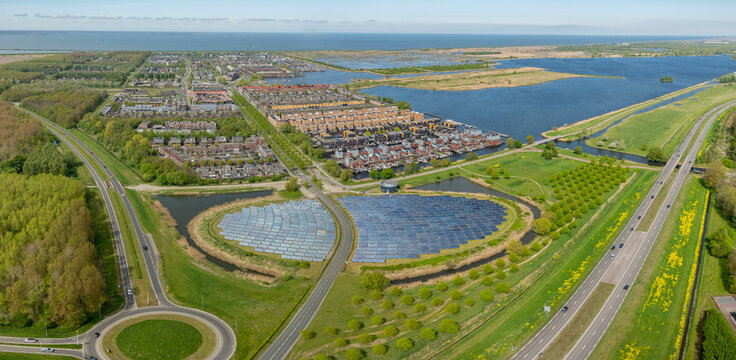Aerial view of solar power plant shaped like island, integrated into green landscape next to modern residential area, featuring solar panels, pond, and bus lane. City heating powered by solar panel.