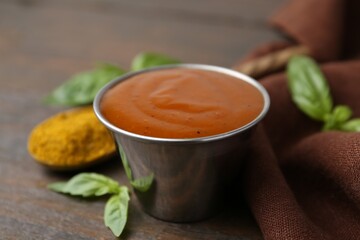 Tasty curry sauce in metal dish, powder and basil on wooden table, closeup