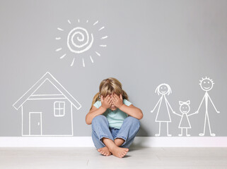 Orphanage. Sad little girl on floor near grey wall with drawings of house, family and sun