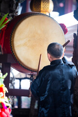 a person dressed in dark traditional attire is intently beating a large temple drum