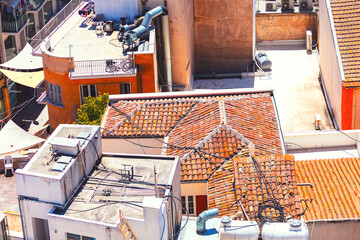 Aerial view shows a cluster of buildings with red-tiled roofs and modern rooftops, adorned with pipes, vents, and equipment. Scene includes a mix of orange and white structures under sunny sky