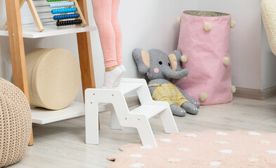 Little girl standing on step stool and reaching for toys on shelf indoors