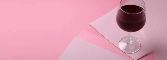 Glass of red liquid with a long stem stands on layered pink surfaces, casting a shadow. The composition features varied shades of pink, creating a sophisticated aesthetic