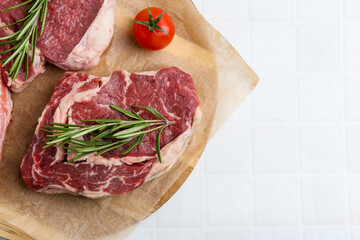 Pieces of raw beef, rosemary and tomato on white tiled table, top view. Space for text