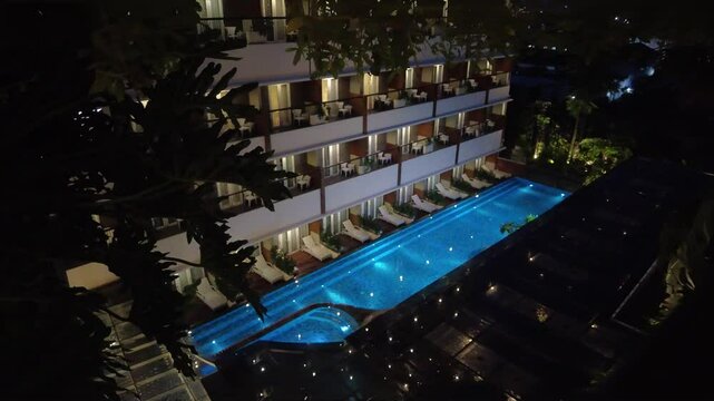 Aerial night view of a hotel pool and building.  Static camera, illuminated pool, tropical setting.