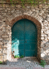 A weathered turquoise door stands in an arched stone entrance, illuminated by soft sunlight. Green vines peek through the cracks in the surrounding textured wall.