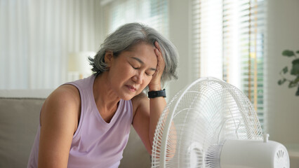 Asia adult people middle age old woman sit at sofa home feel hot flash fever use fan waving hand...