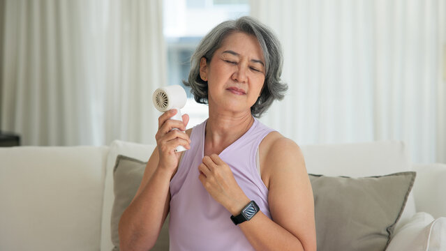 Asia adult people middle age old woman sit at sofa home feel hot flash fever use fan waving hand blowing wind face elder health care summer heat aging issue. Sad pain sweat tired stroke suffer stress.