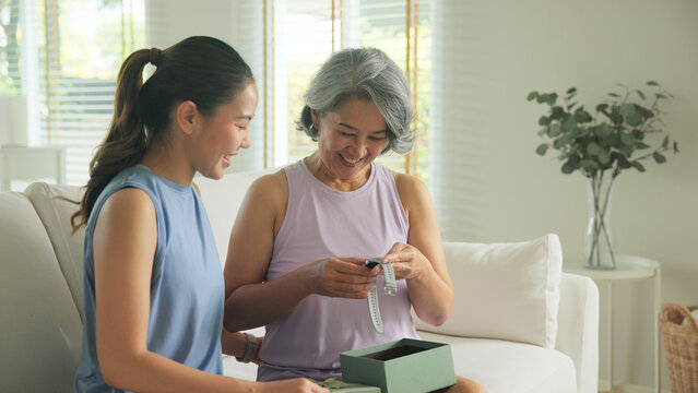 Young adult child give gift AI smart watch IOT app to mature middle age old mum. Love health care older mom asia elder people sit at home sofa happy family wear wrist trace help protect aging life. - Powered by Adobe