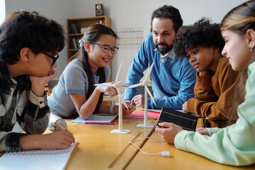Group of mixed-race students engaging in collaborative classroom discussion with teacher, examining wind turbine models and taking notes. Multi-ethnic group of children showing interest while learning