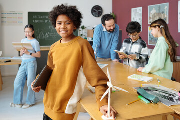 Smiling young students engaged in a group project in classroom setting holding devices and models, with teacher supervising in background