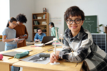 Smiling boy wearing glasses working on small wind turbine project in classroom. Background features classmates and shelves filled with books and school supplies