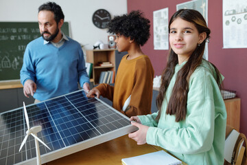 Multiracial students engaging with renewable energy model in classroom setting with teacher explaining solar panels and wind turbines