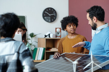 Fototapeta premium Group of diverse individuals discussing renewable energy models in classroom setting with teacher showing solar panel and various educational materials visible in background