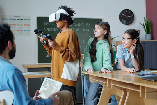 Students engaged in virtual reality experience in classroom, using VR headsets and controllers, while interacting with teacher and peers, fostering interactive and immersive learning environment
