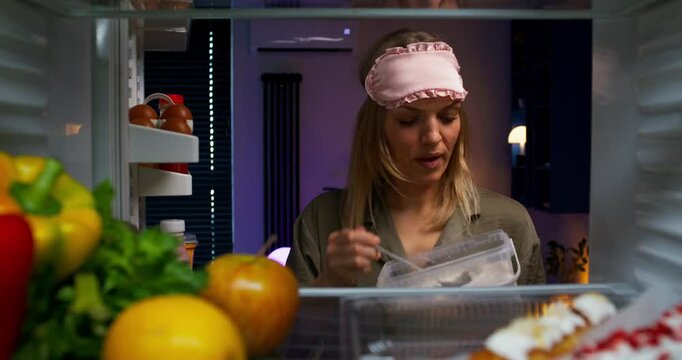 A woman with a sleep bandage on her head eats ice cream from a container while standing near an open refrigerator at night. View from the refrigerator