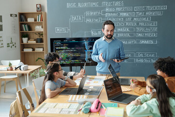 Teacher explaining programming concepts to diverse group of children in classroom setting with visible codes on screen and blackboard. Students paying attention, interacting, using laptops