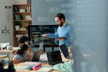 Teacher explaining coding concepts to attentive students in classroom filled with laptops and...