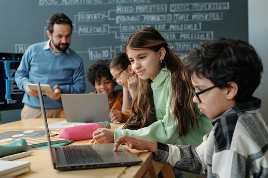 Group of diverse students collaborating on laptop projects with teacher guiding in classroom setting. Background includes chalkboard with equations written - Powered by Adobe