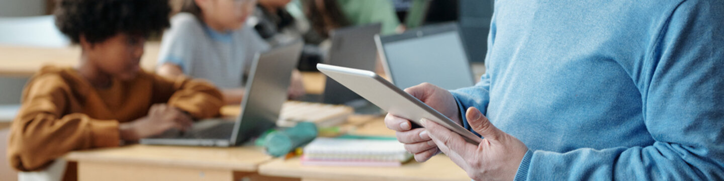 Image showing teacher holding tablet while conducting lesson for students in classroom. Several students are seen working on laptops in background creating collaborative learning environment