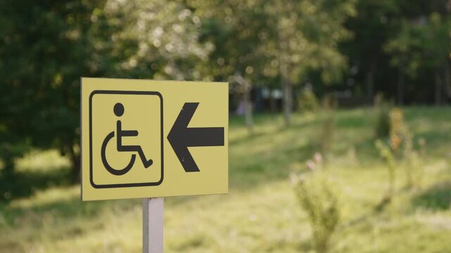 Yellow sign with black wheelchair icon and left arrow signals accessibility. Blurred outdoor backdrop with greenery underscores inclusion in public setting in pakr outdoor on sunny day.