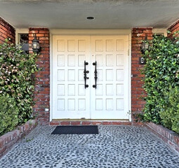 front door of a American house: white wooden double doors with panels; brick wall, stucco ceiling, welcome mat, cobblestone stone walkway, shrubs, hedges, ivy