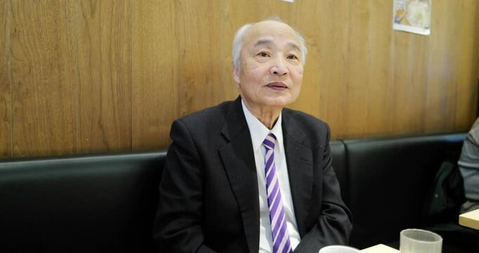 A man in his 70s, wearing a suit, sits at a table in a restaurant and speaks calmly. Surrounded by a peaceful dining atmosphere, he shares his thoughts slowly in a relaxed and comfortable setting.