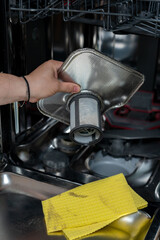 Woman taking out the dirty filter from the dishwasher to clean it on the background of a yellow dishcloth. Concept of cleaning the dishwasher, care and maintenance of kitchen appliances.
