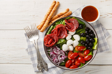 Pizza bowl salad with salami sausages, mozzarella cheese, tomatoes, olives, onions, romaine lettuce and spicy dressing served with breadsticks close-up. Horizontal top view from above