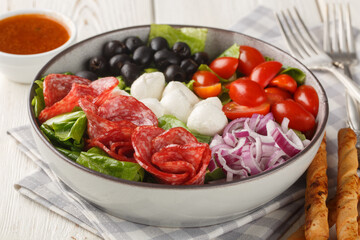 Italian fresh pizza salad with sausages, mozzarella cheese, tomatoes, olives, onions, romaine lettuce and spicy vinaigrette dressing served with breadsticks close-up in a bowl on a table. Horizontal
