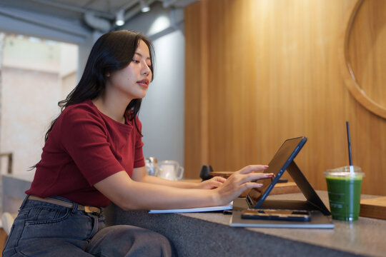 Young asian woman using tablet and working remotely in modern cafe with wooden wall, enjoying a healthy green juice while typing on digital device
