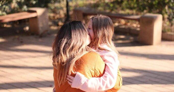 Tender moment between woman and young girl embracing on sidewalk. Mother kisses child standing close suggesting loving relationship on sunny day