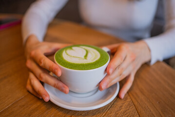 Woman's hands cradling a cup of matcha latte adorned with intricate latte art, savoring a tranquil moment in a cozy cafe with a warm wooden table