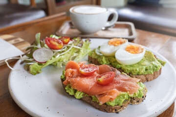Delicious toasts with avocado, smoked salmon, eggs and a variety of fresh vegetables are served on a rustic plate. A healthy and hearty breakfast in an outdoor cafe.