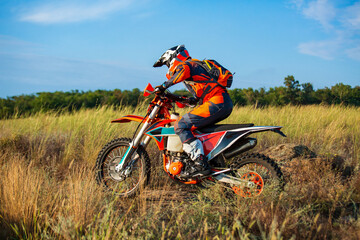 A motocross rider in bright orange gear riding an off-road motorcycle through tall golden grass under a blue sky. Action sports adventure in the countryside.