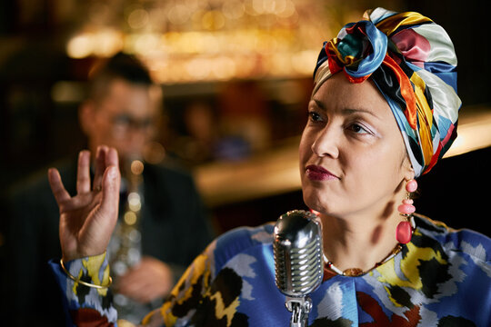 Close up on multiethnic female singer in colorful headwrap passionately performing music with jazz band during live concert at bar, copy space