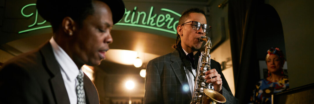 Website header shot of multiracial male musician in suit playing saxophone during jam session with jazz band in bar