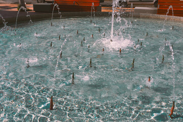 Circular fountain with multiple jets sprays water upward, creating splashes and ripples in shallow pool. Red benches and paved walkway surround fountain, adding vibrant contrast to water blue tones