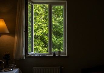 Fototapeta premium A room with an open window showing green trees and a lamp on a table near a curtain and glasses