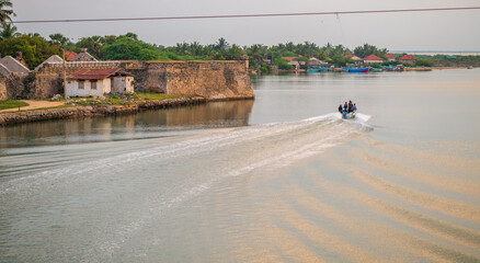 Obraz premium A small fisherman boat speeds across the calm waters near the historic Mannar Dutch Fort in Sri Lanka, leaving a rippling trail behind under a golden sunset sky