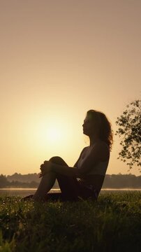 A serene and graceful silhouette of a woman sits peacefully by the tranquil waterside during a stunning sunset, embodying a sense of tranquility and deep introspection amid the beauty of nature