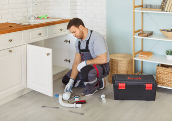 Professional male plumber in uniform with toolbox sitting near sink replacing the water filter or fixing broken pipe in modern kitchen. Repair service and plumbing maintenance concept.