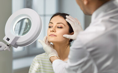 Beautiful cheerful young lady sitting in beauty clinic and having consultation about plastic surgery with a male doctor cosmetologist. Surgeon hands touching woman face examining skin.