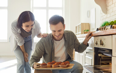 Portrait of young happy couple looking at baked muffins on baking tray standing in kitchen....