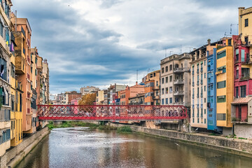The iconic red iron bridge Pont de Ferro by Gustave Eiffel, spans the Onyar River in Girona, Spain, framed by colorful houses and an overcast sky. A birds fly around