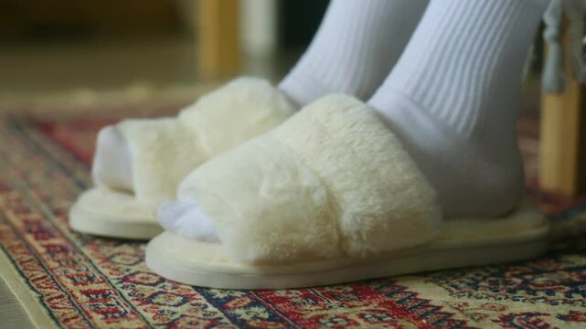 Soft white slippers wait on plush carpet. Woman in warm white socks puts them on before starting her winter day. The shot conveys warmth and protection from the cold. Home comfort in winter day.