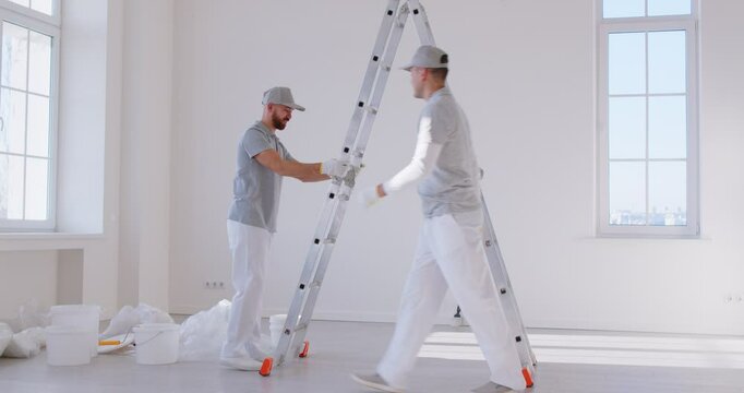 Two professional male painters discuss painting wall in spacious, bright room. Men in gray work uniforms bring ladder into room, where while various painting supplies are scattered around floor.