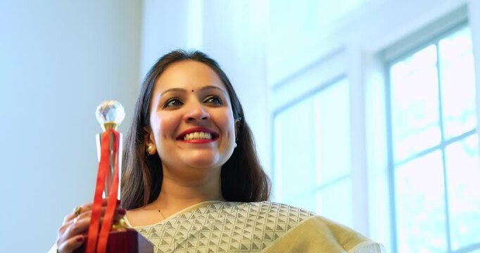 Holding trophy with pride, Indian young successful female in traditional saree says thank you to audience while receiving award on stage for exceptional work, captured from low angle in closeup view