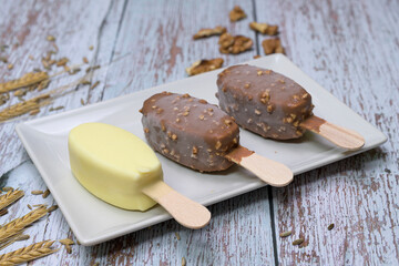 Delicious scene with three ice cream bars, including chocolate and vanilla flavors, served on a white plate with toppings, seeds, nuts, and colorful sprinkles. Rustic wooden background